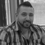 A man with short dark hair and a beard, wearing a plaid shirt, sits indoors and smiles at the camera.