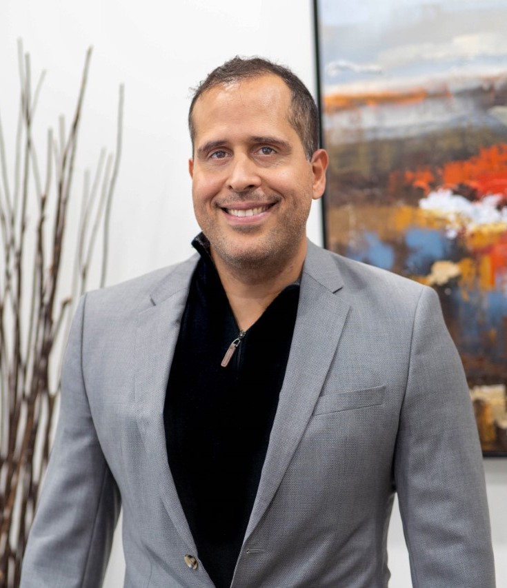 A man in a grey suit jacket and black shirt stands indoors, smiling at the camera, with abstract artwork and decorative sticks in the background of an architecture company.