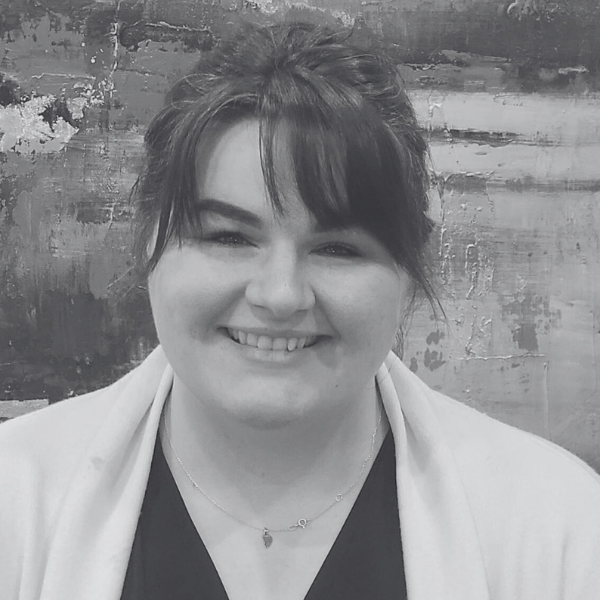 A woman with dark hair and bangs smiles at the camera, wearing a light-colored cardigan and a necklace, in front of an abstract painted background at an architecture company.
