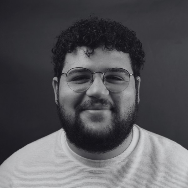 A person with curly hair, a beard, and glasses is smiling at the camera, wearing a light sweater. The plain, dark background gives a professional look—perfect for an architecture company profile photo.