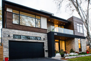 Modern two-story house with wood and stone exterior, large windows, black garage door, balcony, and landscaped entrance with potted plants. House number 384 is visible.