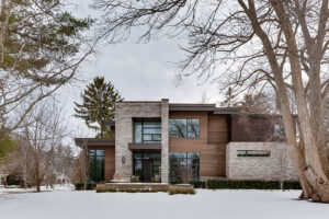 A modern two-story house with large windows and a mix of wood and stone exterior, surrounded by trees and snow-covered ground on a cloudy day.