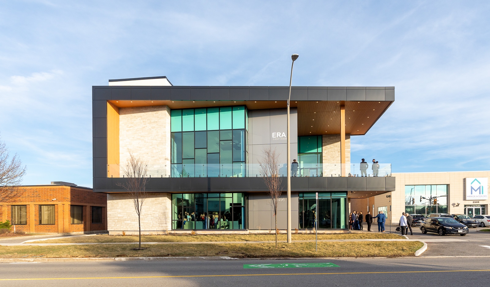 A modern two-story commercial building with glass windows and a cantilevered upper level. Several people are gathered near the entrance and on the upper balcony.