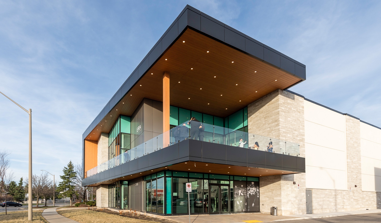 A modern two-story commercial building with large glass windows, a cantilevered roof, and a glass balcony, set on a landscaped street corner.