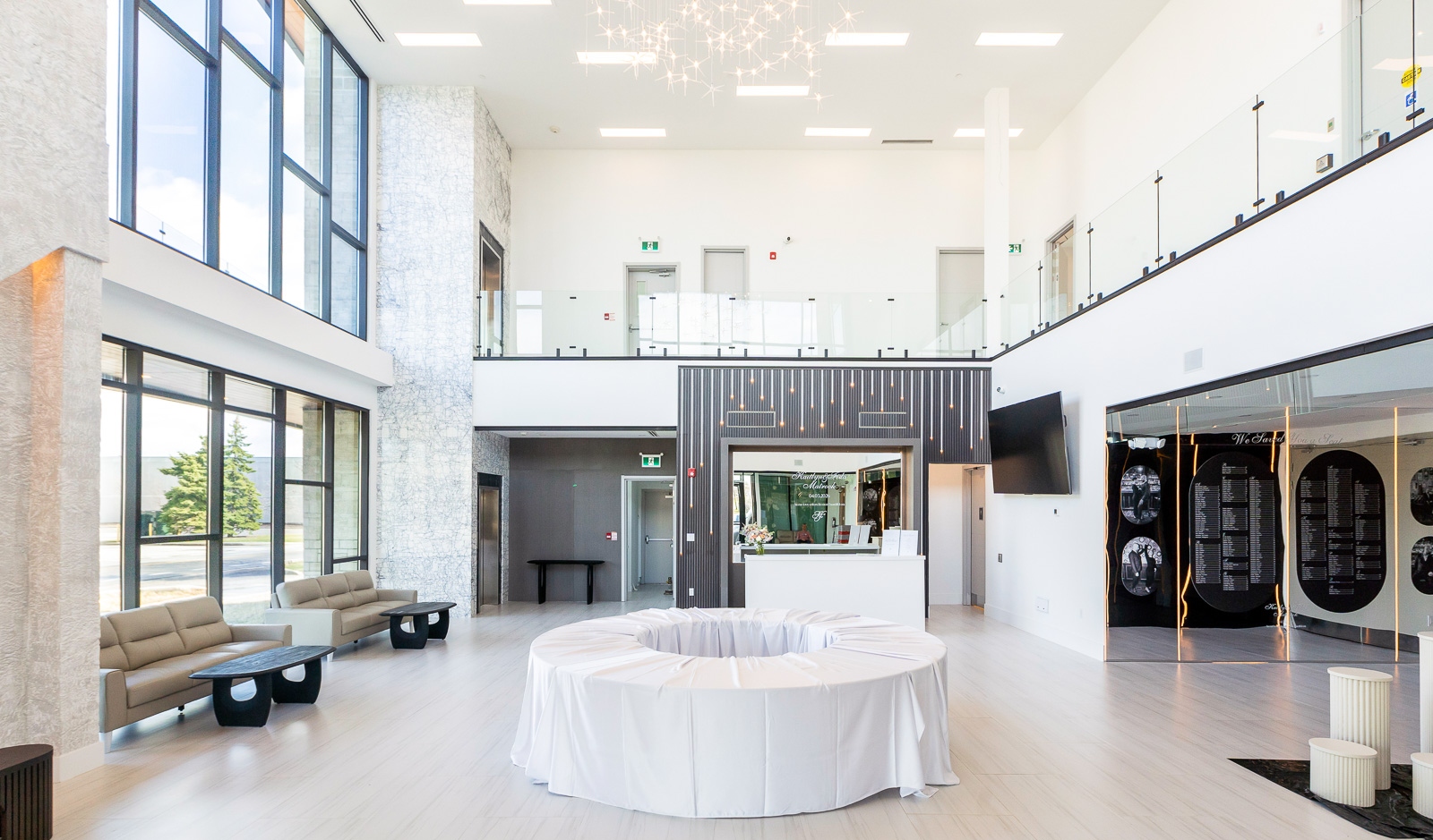 Spacious, modern lobby with high ceilings, large windows, beige seating, a round draped table in the center, and a reception desk near the back wall.
