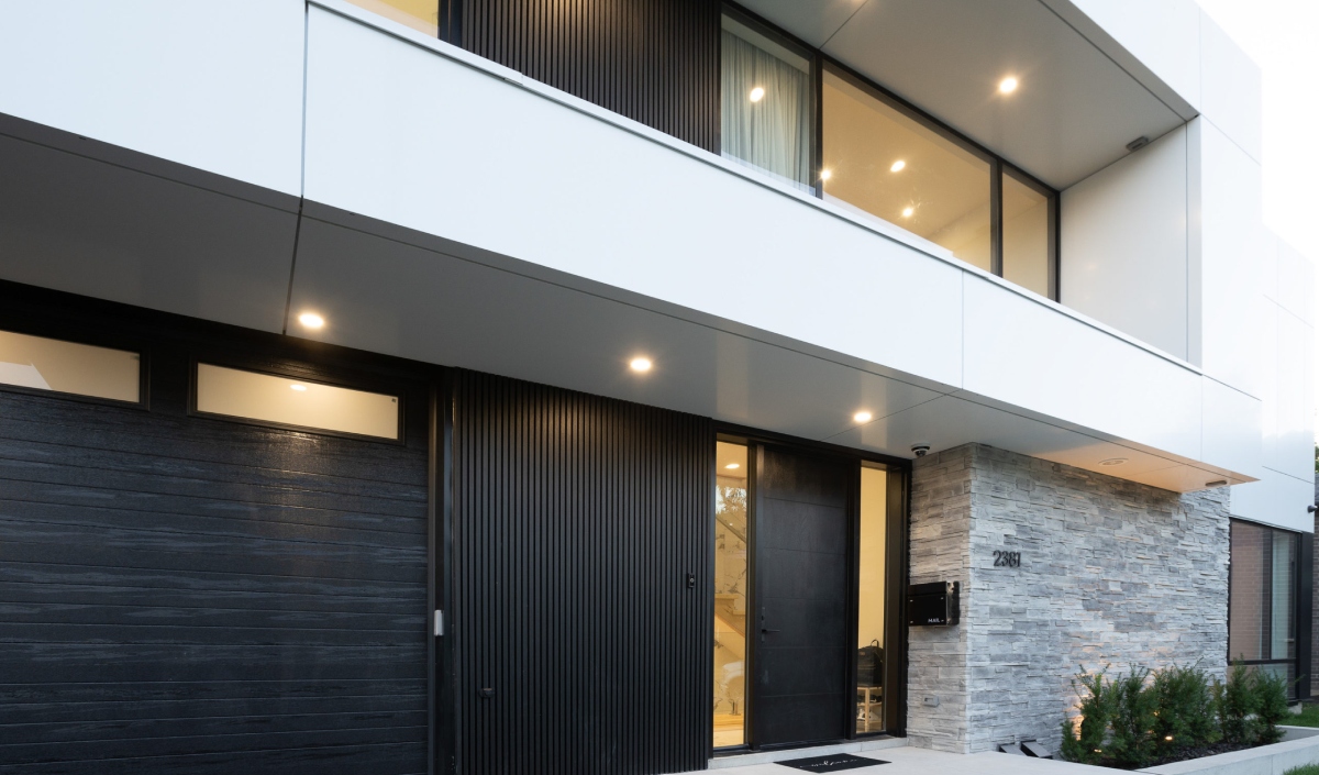 Modern custom home exterior with large windows, black panel doors, a stone accent wall, and warm recessed lighting at the entrance.