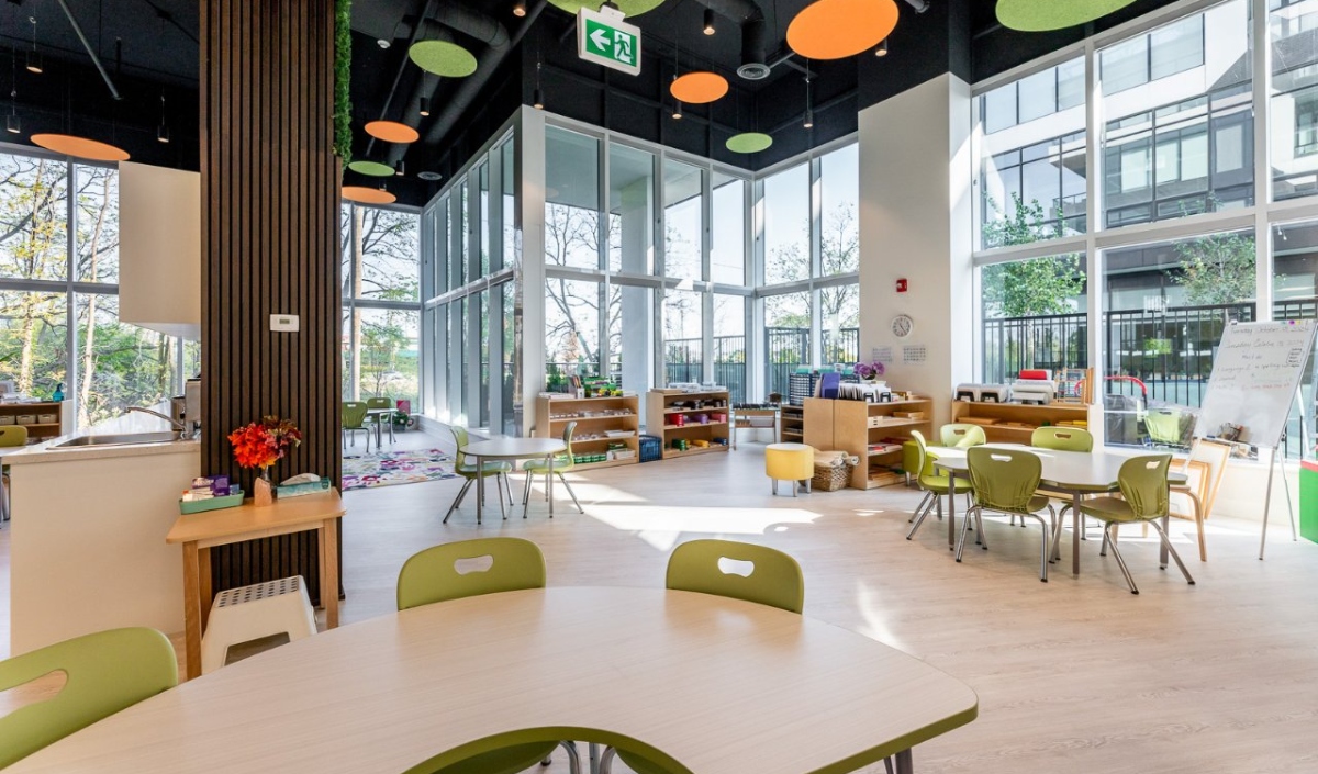 Bright, modern classroom with large windows, green chairs around tables, shelves with books and toys, and colorful circular ceiling decorations.