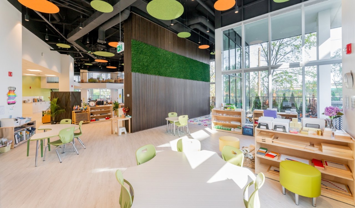 Bright, modern classroom with round tables, green chairs, open shelves, large windows, and colorful ceiling panels. Natural light fills the space, highlighting educational materials.