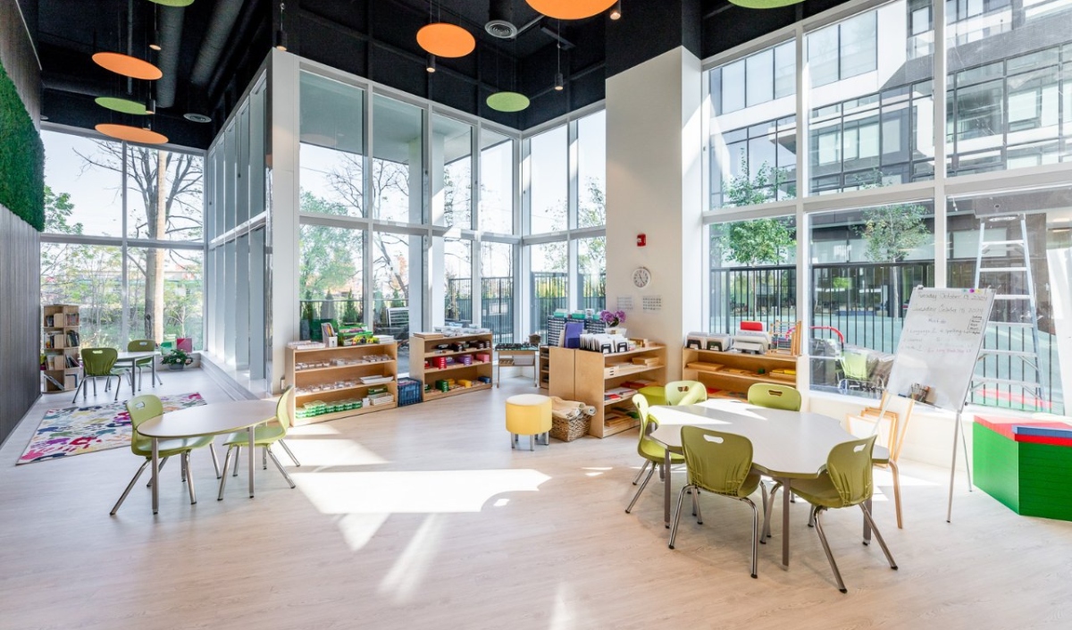 A bright, modern classroom with large windows, several tables and chairs, open shelves with educational materials, and colorful, circular ceiling decorations.