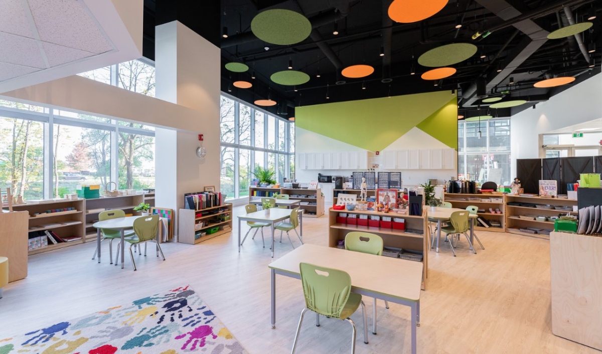 A modern classroom with large windows, several tables and green chairs, bookshelves, colorful ceiling panels, and a rug with handprints.