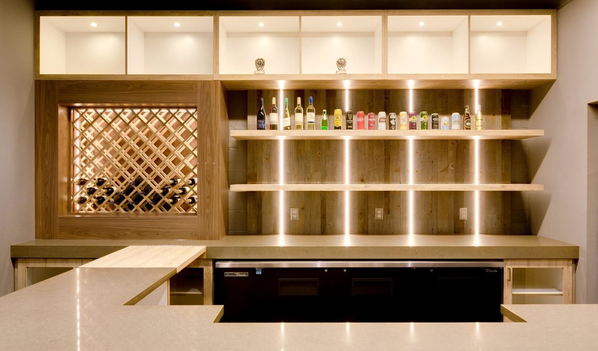 Modern bar area with built-in wine rack, bottles displayed on illuminated wooden shelves, and a light-colored countertop.