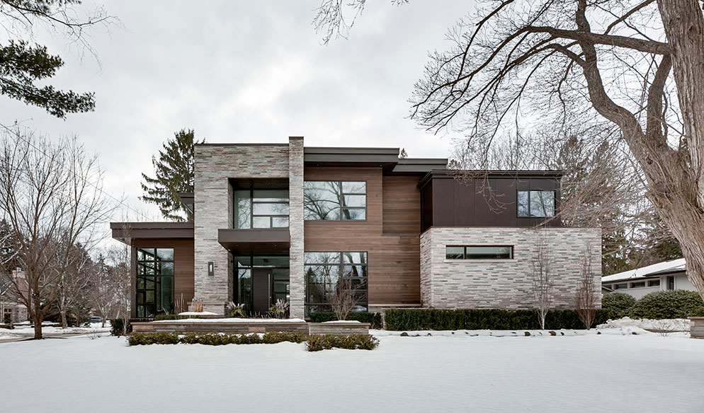 Modern two-story house with large windows, stone and wood exterior, surrounded by leafless trees and snow-covered ground.