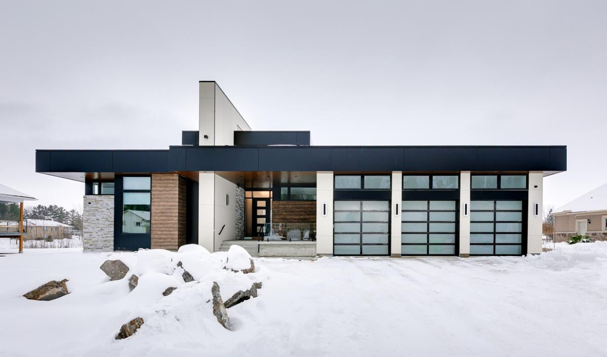 Modern house with flat roof, large glass garage doors, and wood and stone accents, surrounded by snow and rocks under an overcast sky.