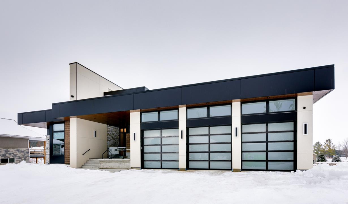 Modern house with flat roof, large glass garage doors, and beige exterior walls, surrounded by snow on the ground.