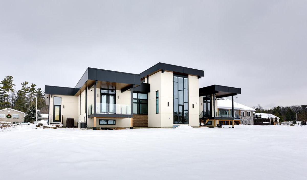 Modern house with large windows and flat roofs stands on a snowy landscape under an overcast sky; trees and other buildings are visible in the background.