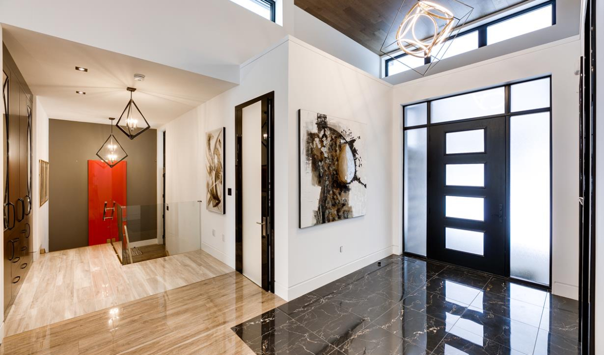 Modern entryway with black front door, marble and wood flooring, geometric light fixtures, and abstract wall art. Stairs with glass railing lead to a lower level at the hallway's end.