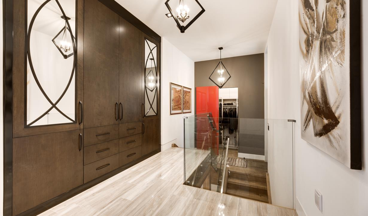 A modern hallway with large dark wood wardrobes, glass railing, light wood flooring, pendant lights, and abstract artwork on the wall.