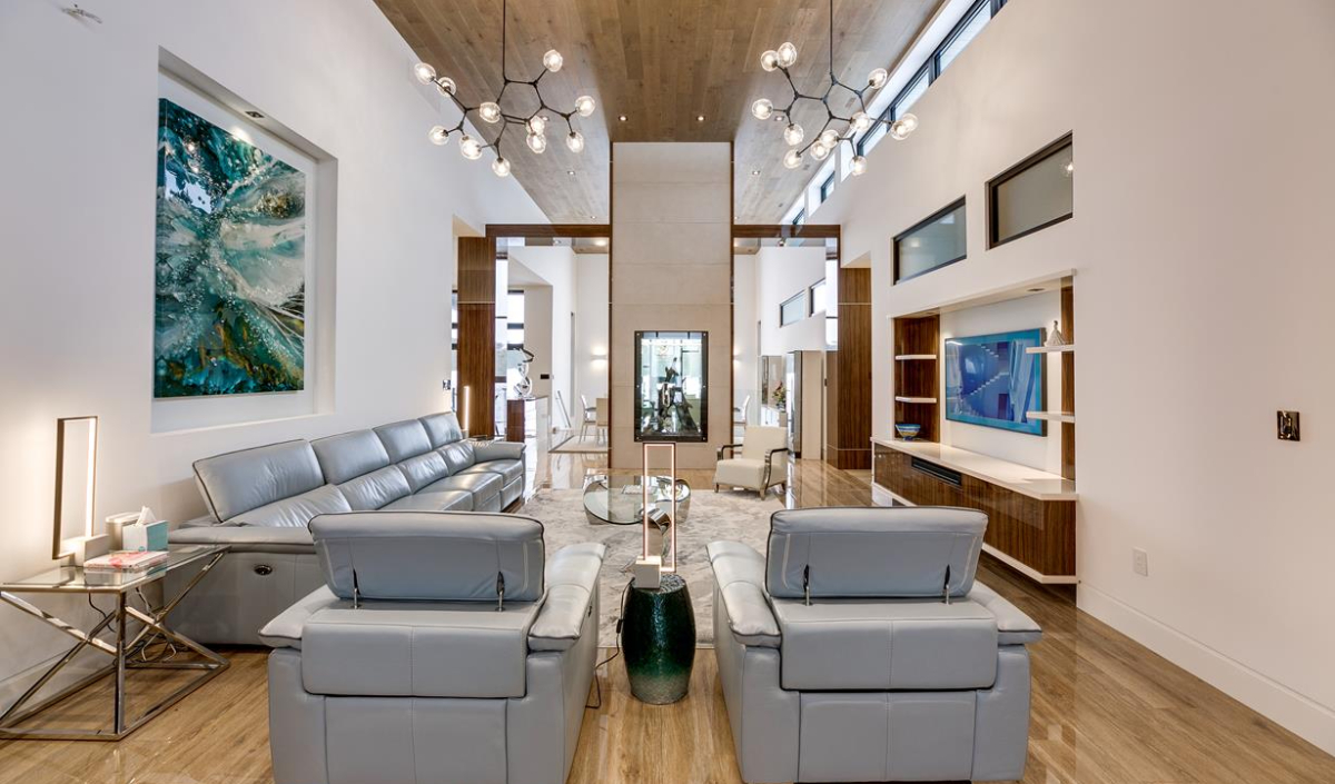 Modern living room with light gray leather sofas, wood ceiling, abstract artwork, and a large chandelier, featuring open shelves and a wall-mounted TV.