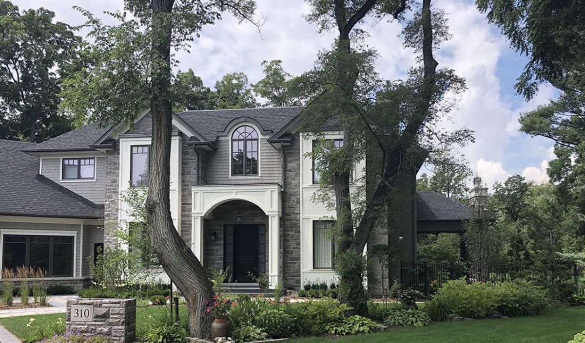 Two-story house with arched entryway, gray exterior, and dark roof, surrounded by trees, greenery, and a lawn. House number 310 is visible on a stone block in the foreground.
