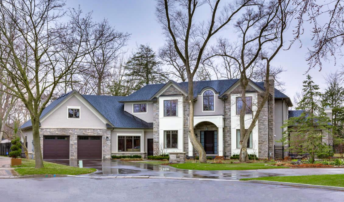 Two-story suburban house with a stone and siding exterior, double garage, large windows, and a curved driveway bordered by trees and wet pavement.