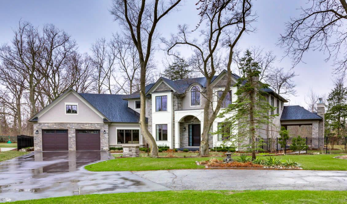 Two-story suburban house with gray siding, stone accents, and a double garage, surrounded by grass, trees, and a wet driveway on an overcast day.
