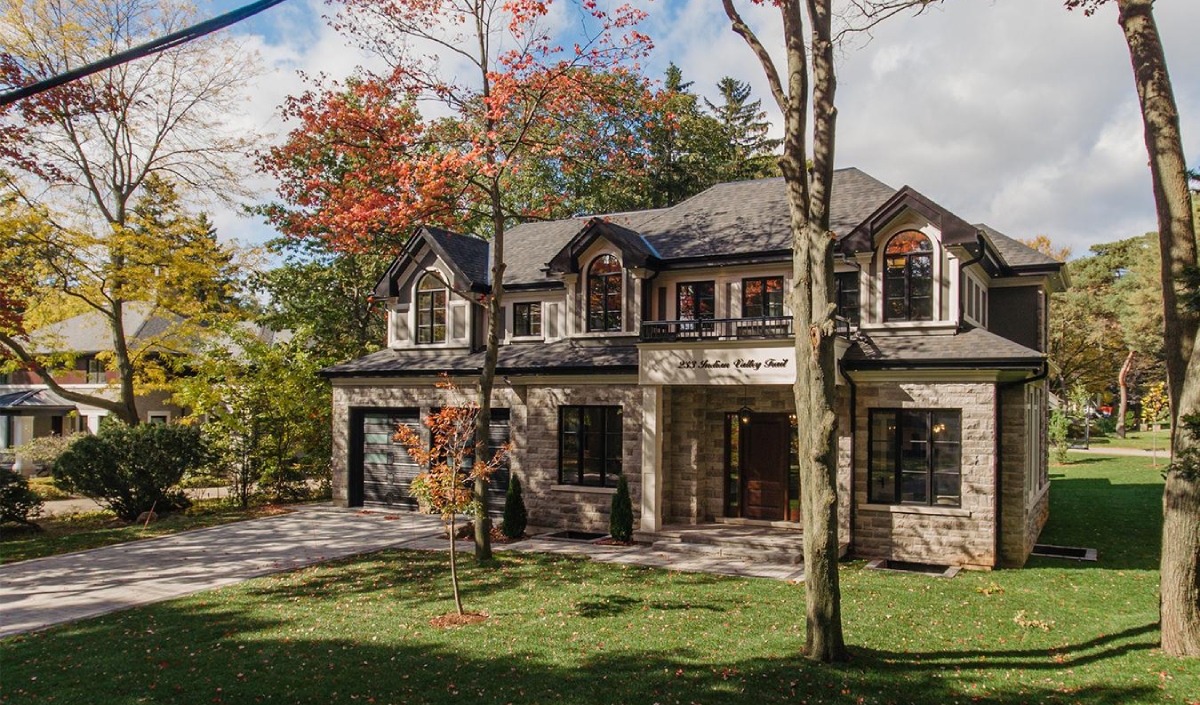 Two-story modern stone house with a dark roof, double garage, driveway, front lawn, and trees with autumn foliage.