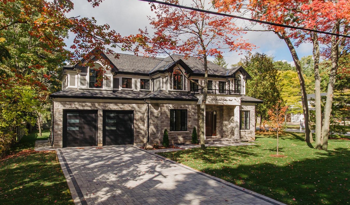 Two-story suburban house with brick exterior, double garage, and paved driveway, surrounded by trees with autumn foliage.