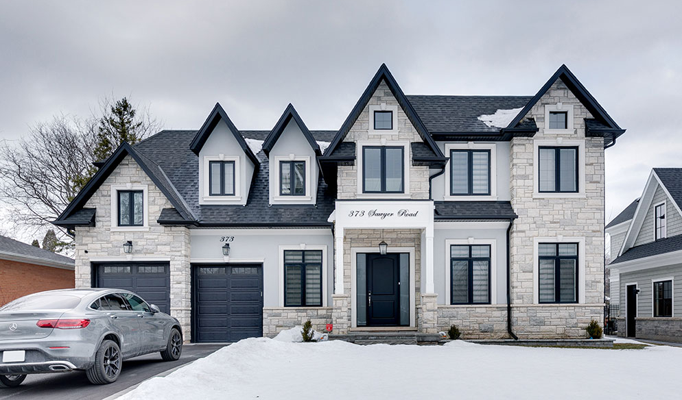 Two-story stone house with black roof and trim, three dormer windows, double garage, driveway with parked car, and snow on the ground.