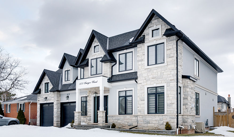 Two-story modern stone house with large windows, three black garage doors, and a covered entrance, surrounded by snow under a cloudy sky.