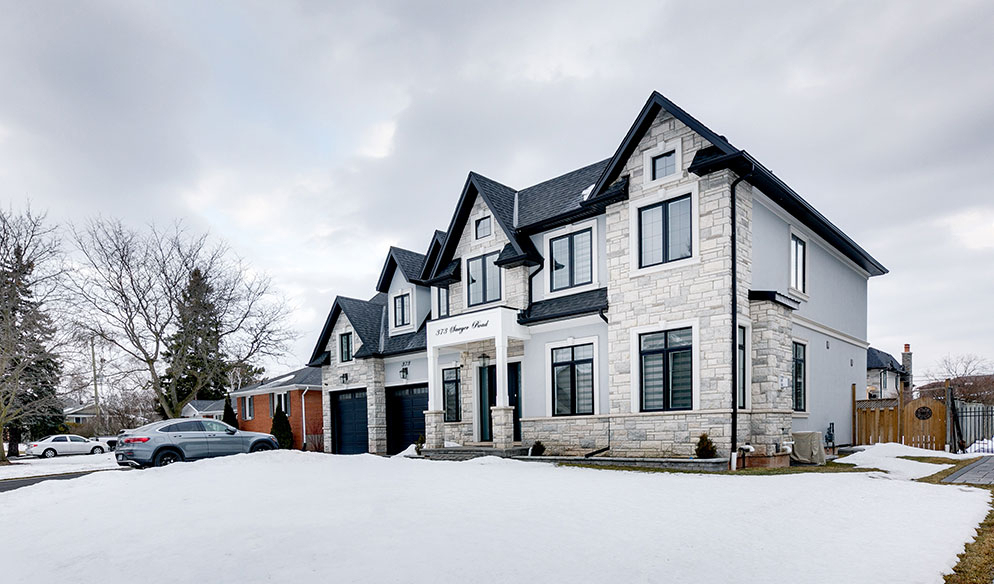 Two-story modern house with gray stone facade, black roof, and attached garage, surrounded by snow with a parked car in driveway under a cloudy sky.