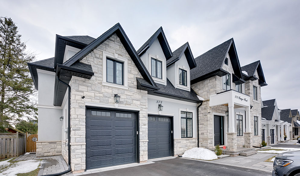 A modern duplex house with two driveways, stone and stucco exterior, black garage doors, and snow patches on the ground.