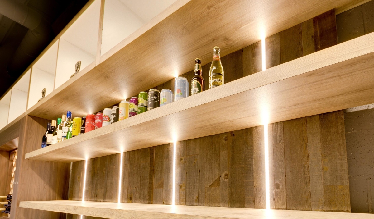 Wooden shelves with assorted bottles, cans, and jars displayed in a row, illuminated by vertical strip lights against a wood-paneled wall.