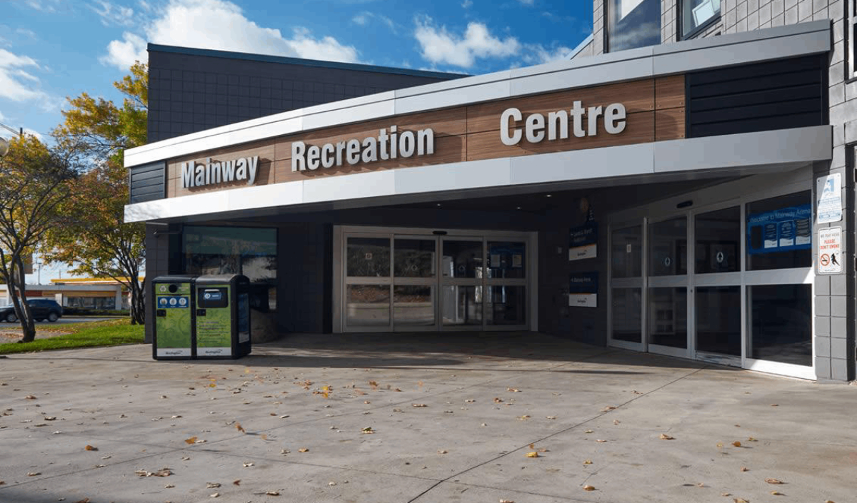 The entrance to Mainway Recreation Centre with automatic glass doors, signage above, trash bins, and scattered fallen leaves on the ground.