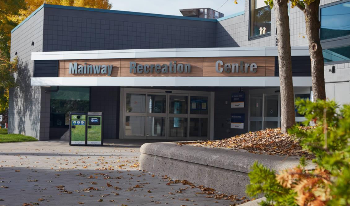 The entrance to Mainway Recreation Centre, with glass doors, signs, and a sidewalk covered in fallen leaves.