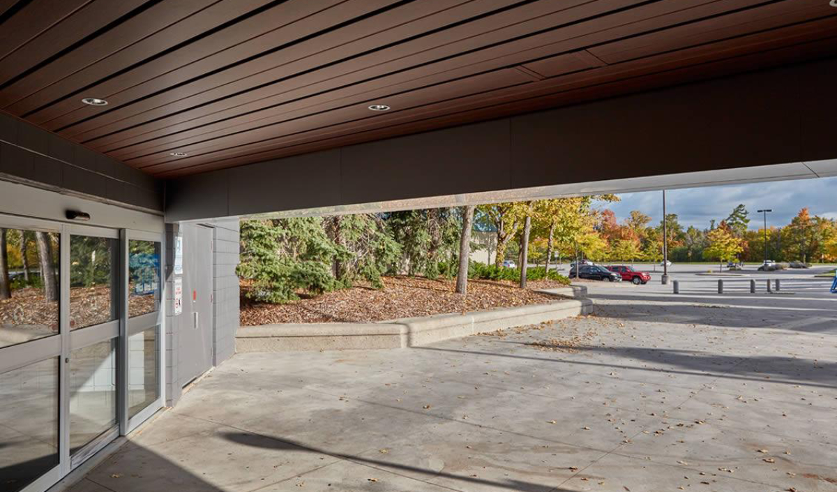 A covered entryway with glass doors opens to a paved area, trees with autumn foliage, and a partially full parking lot under a partly cloudy sky.
