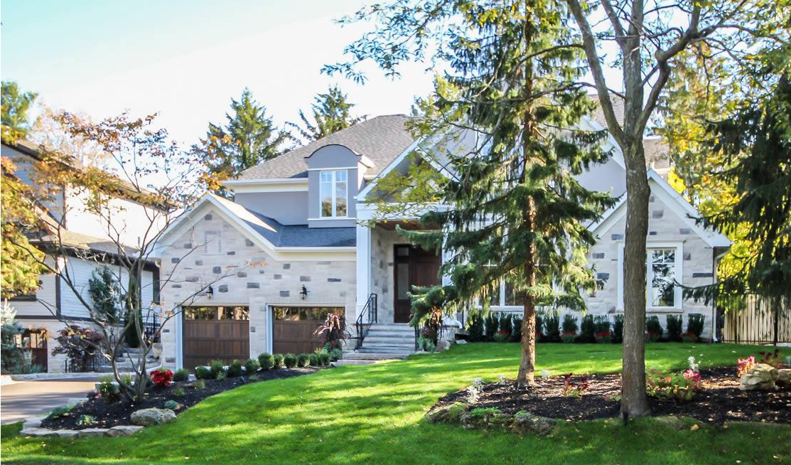 A modern two-story house with a stone facade, two-car garage, front steps, and landscaped lawn surrounded by trees and shrubs.