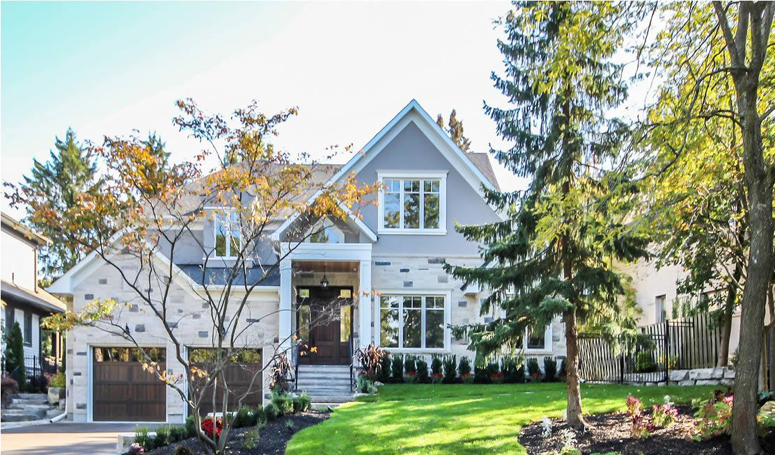 Two-story suburban house with a gray exterior, stone accents, attached garage, manicured lawn, and trees in the front yard on a sunny day.