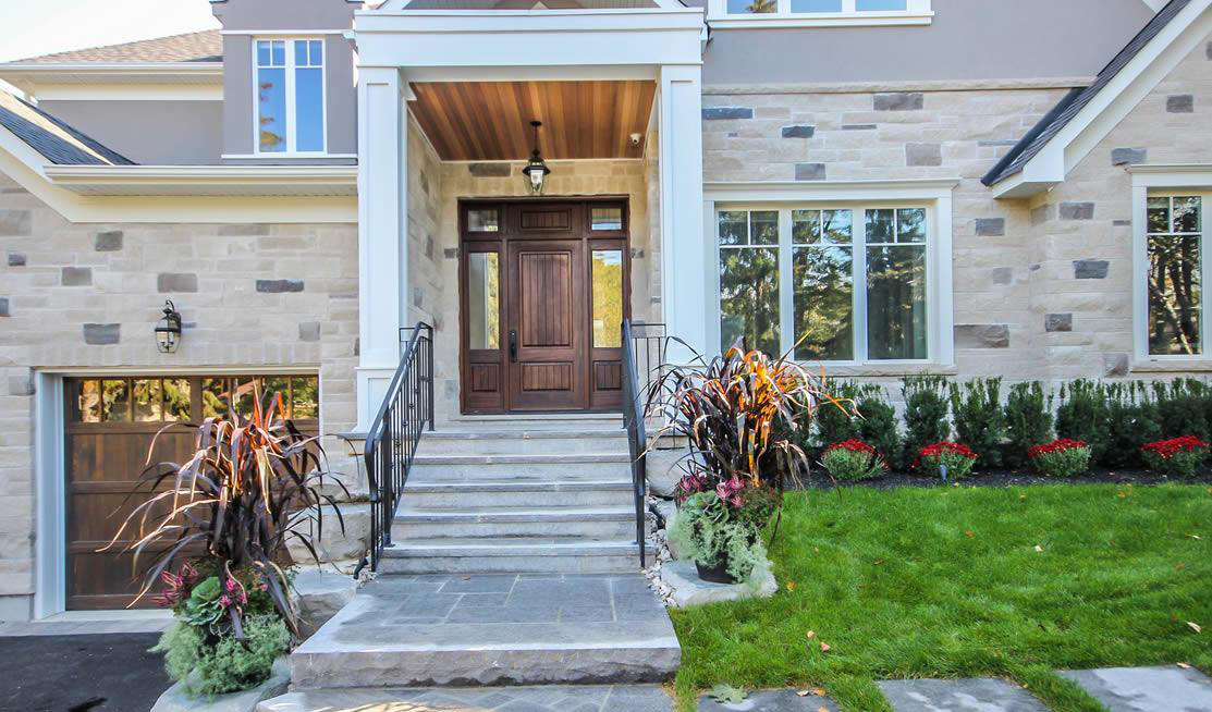 Stone steps with metal railings lead to a wooden front door of a house with stone and siding exterior, manicured lawn, and decorative plants.
