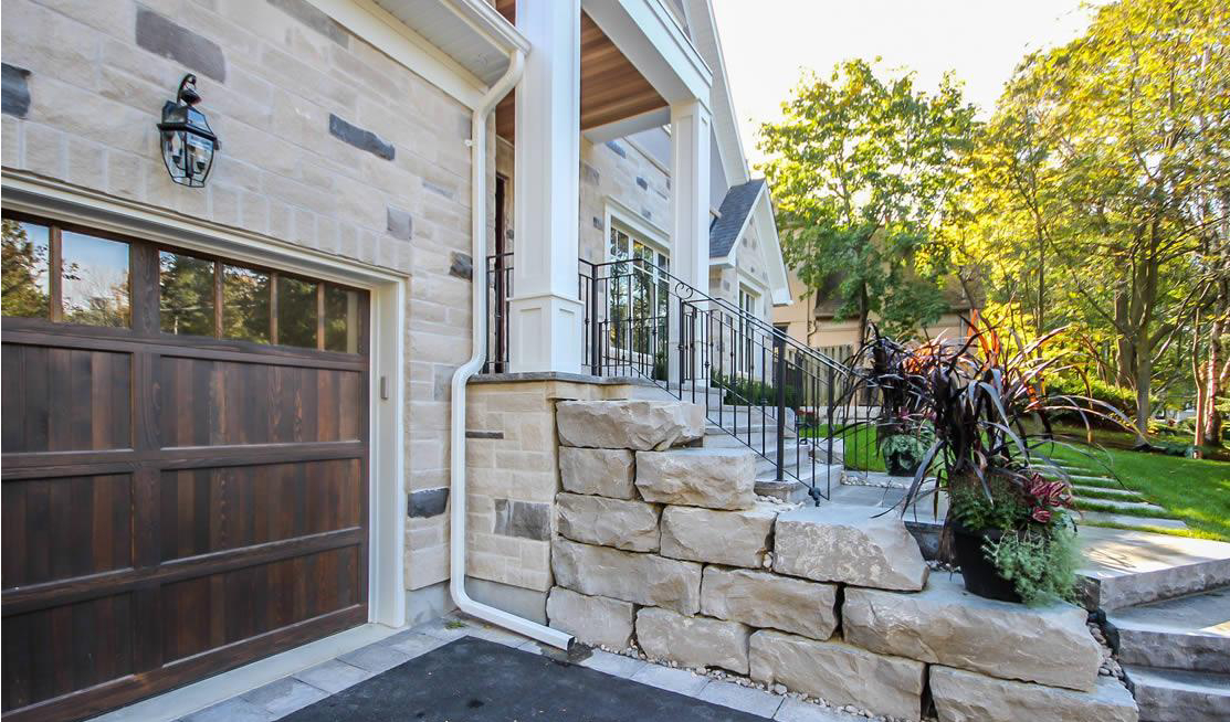 Stone steps lead to a front porch with black railings, next to a garage with a wooden door and landscaped greenery along the walkway.