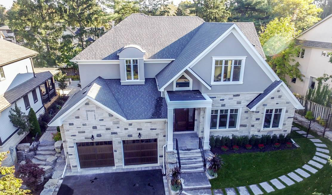 A two-story modern suburban house with a stone and gray exterior, double garage, large front windows, and landscaped front yard.