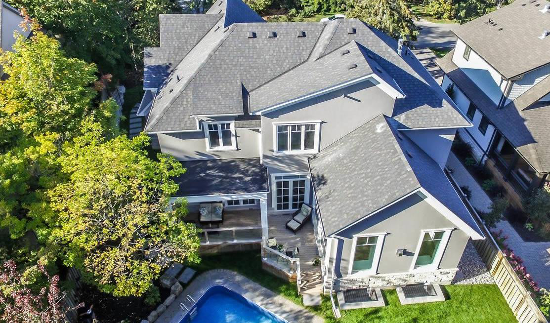 Aerial view of a modern two-story house with a gray roof, backyard patio, outdoor seating area, trees, and a swimming pool.