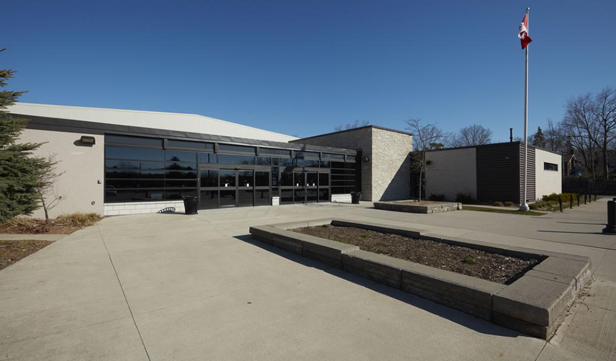 A modern, single-story public building with large glass windows, a flagpole with a Canadian flag, and empty raised garden beds in front under a clear blue sky.