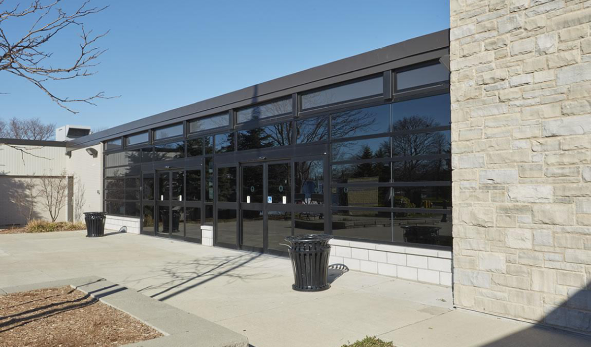 A modern building with large glass windows and stone walls, featuring two black trash cans on a concrete walkway under a clear blue sky.