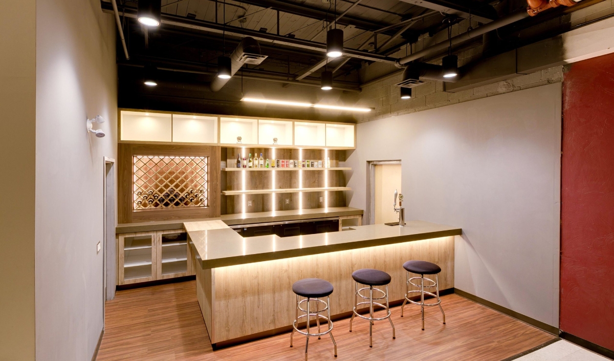 Modern home bar with a wooden counter, three round stools, open shelves with bottles, a wine rack, and overhead lighting.