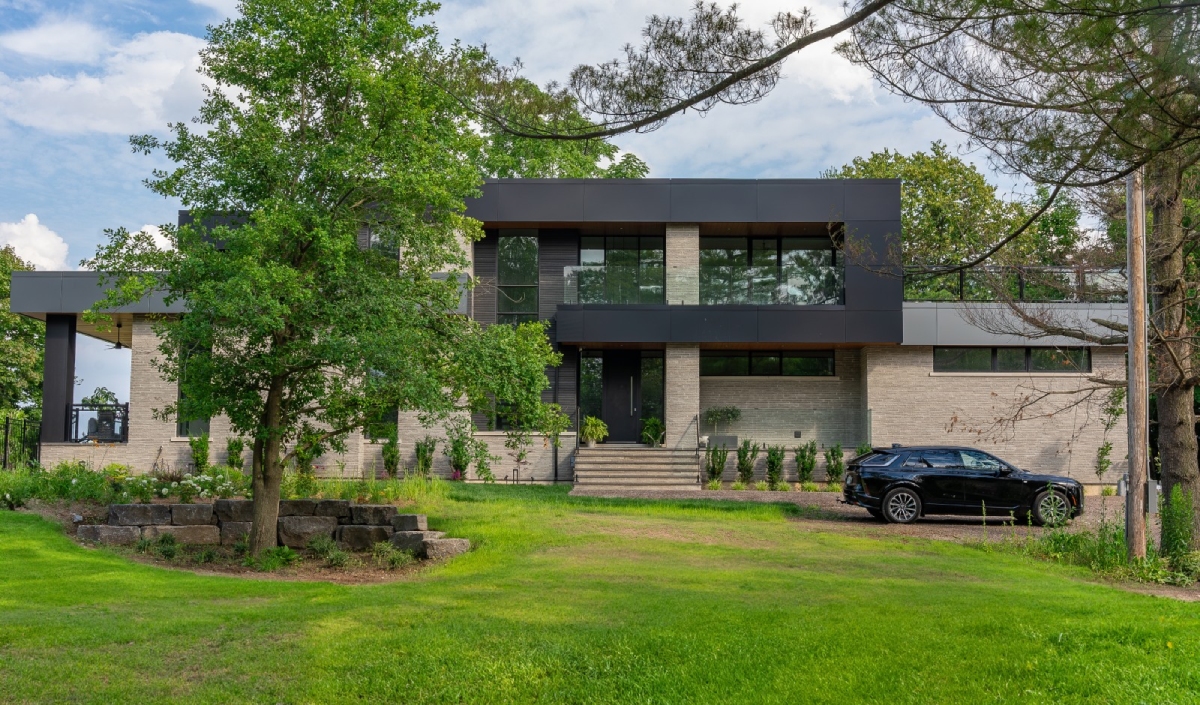 Modern two-story house with large windows, beige brick and dark paneling, surrounded by trees and grass; a black car is parked in the driveway.