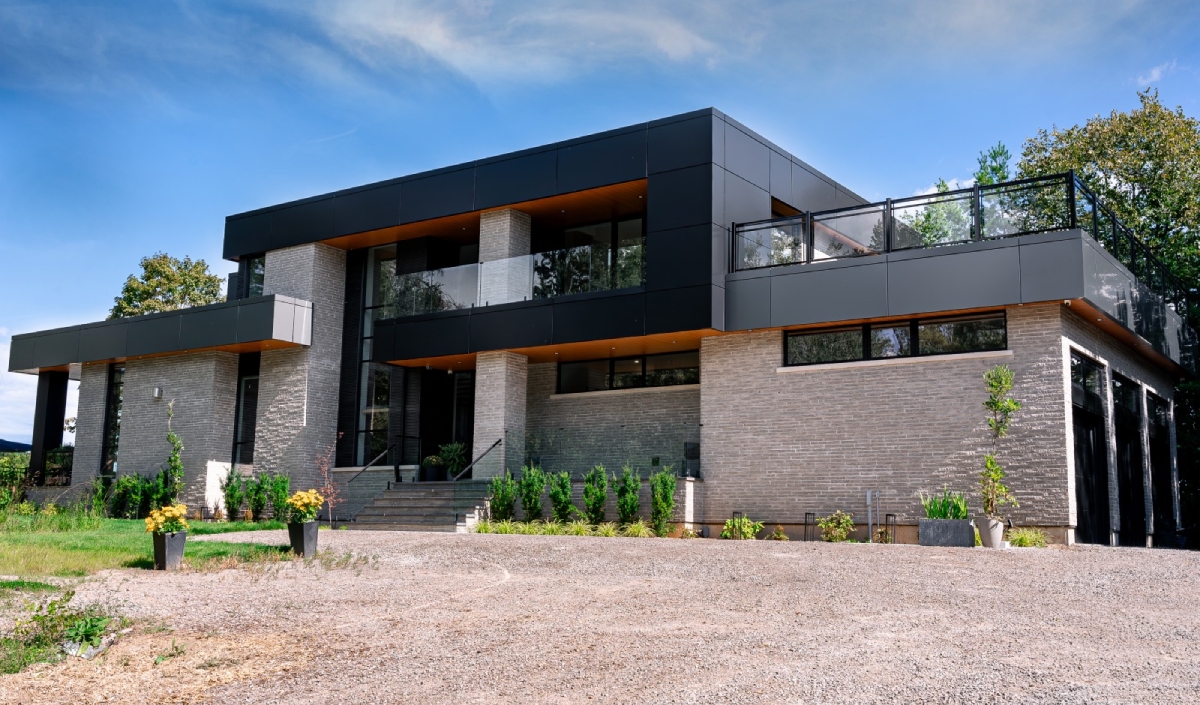 Modern two-story house with dark cladding, large windows, and a brick facade, surrounded by greenery and a gravel driveway under a blue sky.
