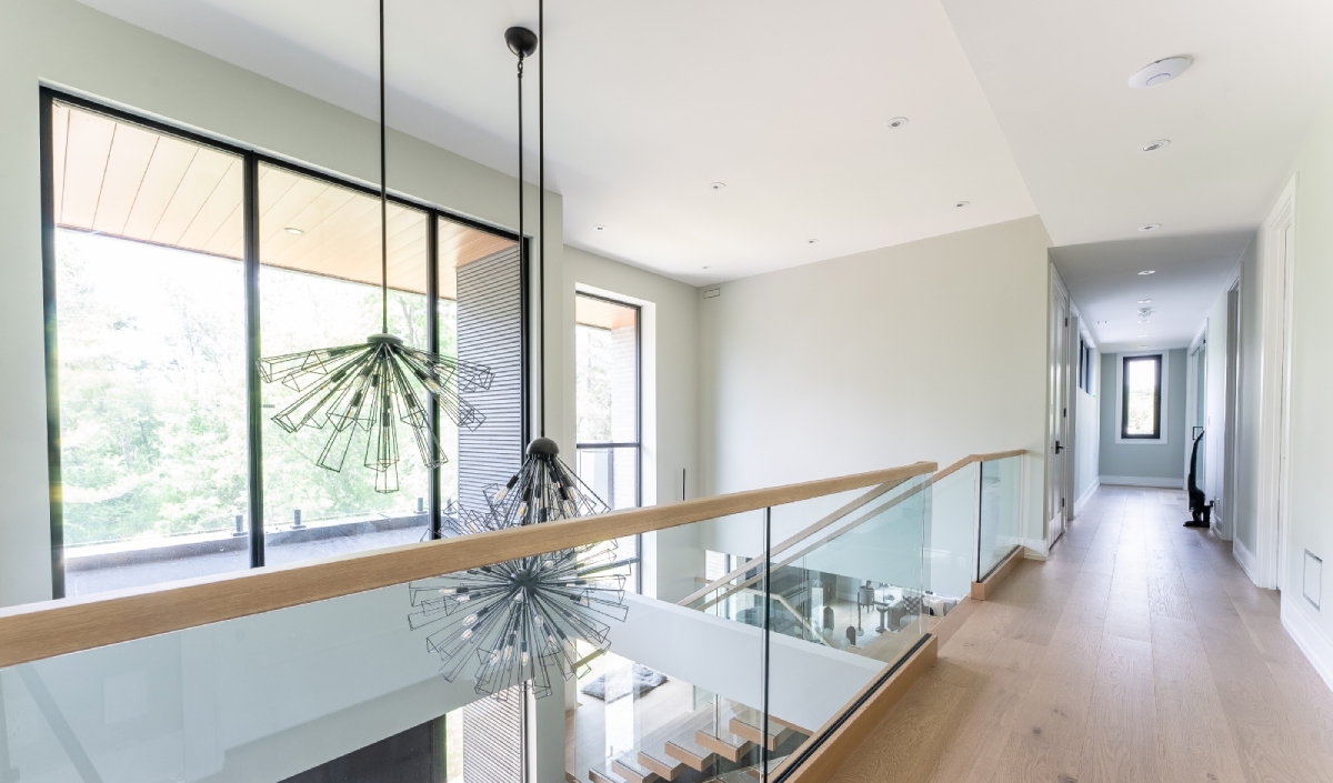 Modern hallway with light wood floors, glass railing, two contemporary pendant lights, large windows, and a staircase leading downstairs.