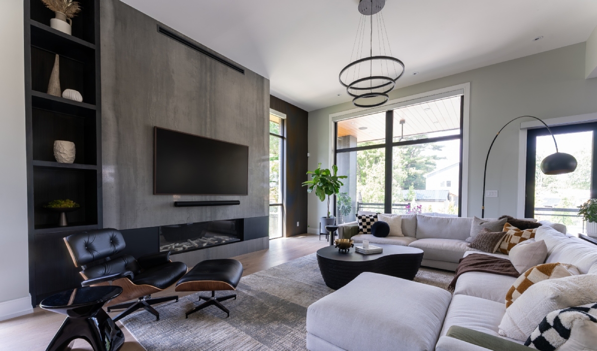 Modern living room with a sectional sofa, lounge chair, large TV on a concrete accent wall, floor-to-ceiling windows, and minimalist decor. Natural light fills the space.