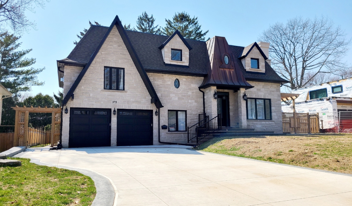 A two-story stone house with steep dark roofs, double garage doors, a copper-toned dormer, steps leading to the entrance, and a curved driveway in front.