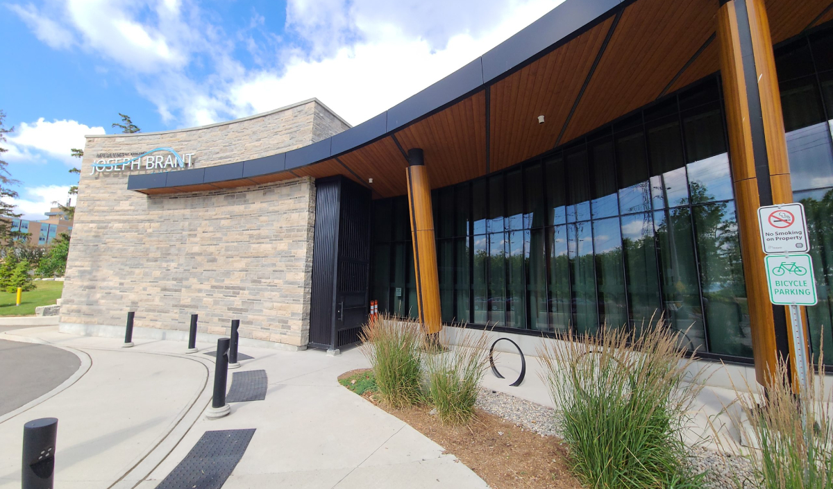 Exterior view of a modern building with "Joseph Brant" signage, glass windows, and stone walls—home to the Joseph Brant Museum—featuring bicycle parking and a no smoking/no vaping sign near the entrance.